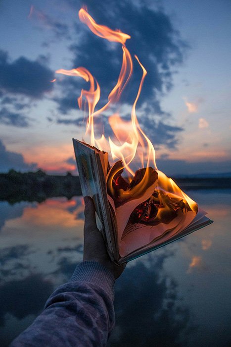 A hand holds a burning book on a lake's shore at sunset
