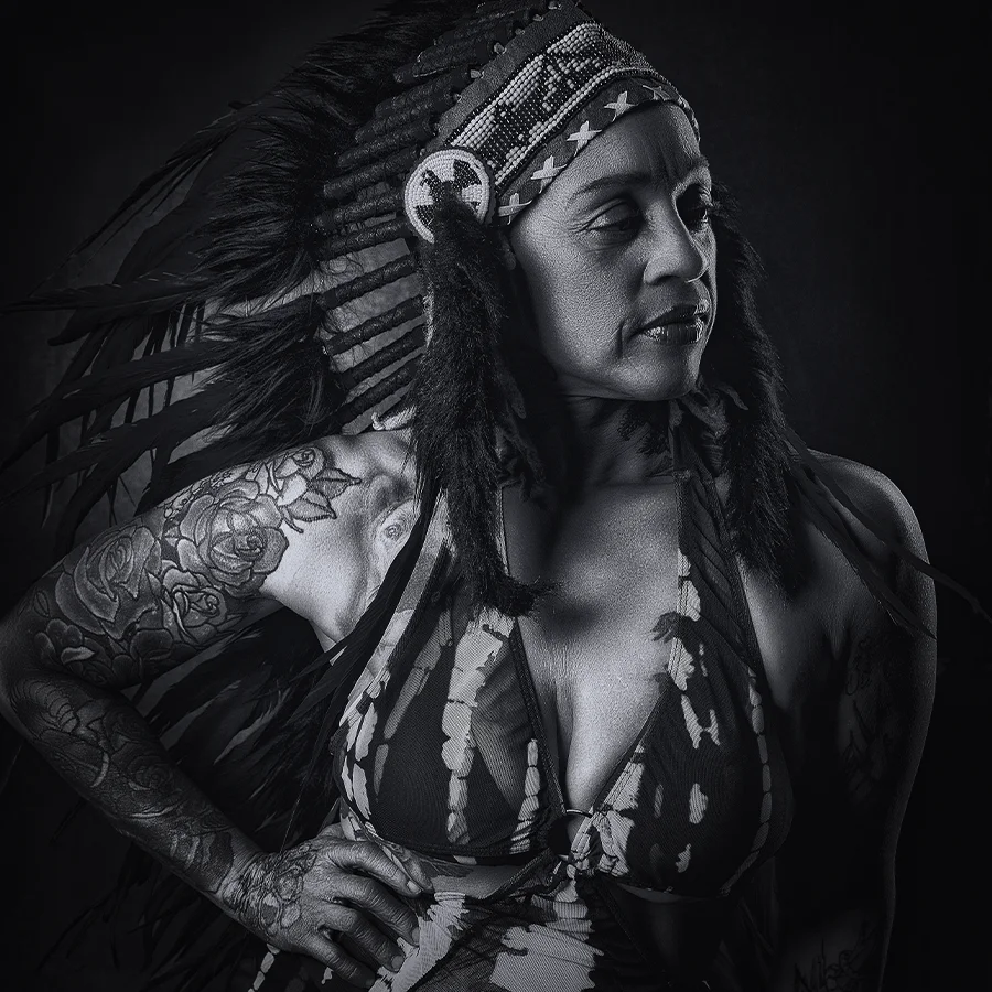 Black and white fine art portrait of a confident woman with tattoos, wearing an elaborate feathered Native American-style headdress and a patterned halter top. She stands in dramatic lighting with her hand on her hip, gazing powerfully to the side. Her left arm features a prominent rose tattoo. Strong, artistic, and striking studio portrait.