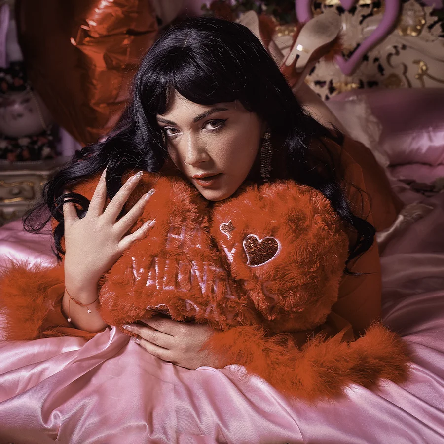 A young lady lies on her back on a red satin bed, she's wearing black and red lingerie and looking up at the camera