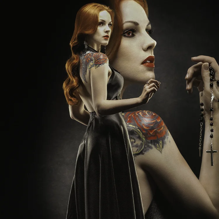 A red head head with shoulder tattoos, wearing an elegant black dress poses beautifully on a textured backdrop. A full headshot image is super imposed on the background, depicting our red headed young lady's face in more detail, you can see her eyes are an ember color, and she's holding a rosary in this vampire style art work.